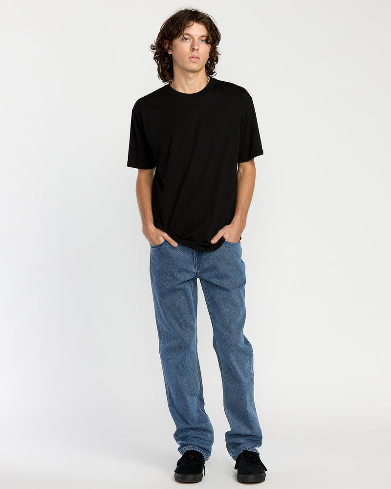 A young person with wavy brown hair wears a plain black T-shirt, Men's Solver Denim in Deep River Blues, and black shoes, standing against a plain white background with hands in pockets.