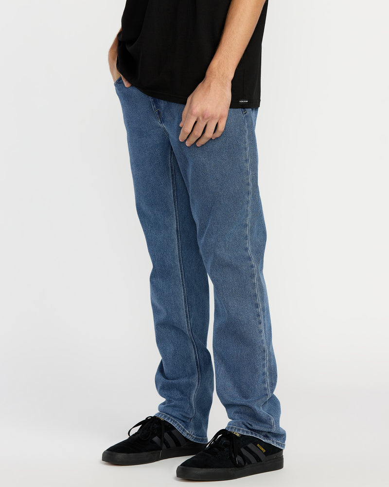 A person wears a black t-shirt, Men's Solver Denim in Deep River Blues by Volcom, and black sneakers with white soles, standing with hands in pockets against a plain white background.