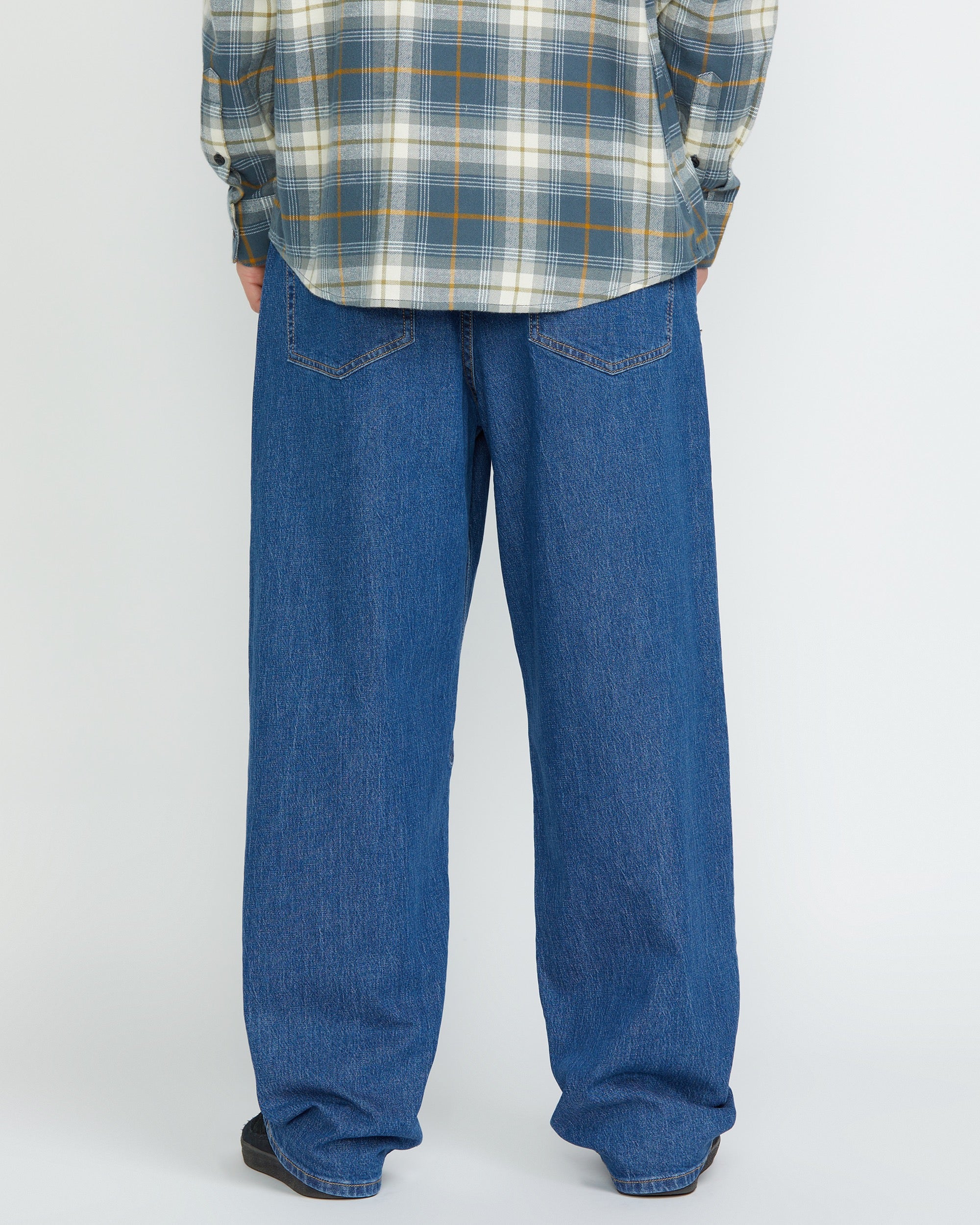 A person stands facing away, showing only the lower half, wearing Volcom Men's Billow Denim Pants and a gray plaid shirt against a plain light background.