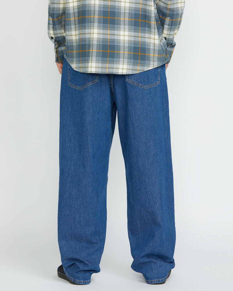 A person stands facing away, showing only the lower half, wearing Volcom Men's Billow Denim Pants and a gray plaid shirt against a plain light background.