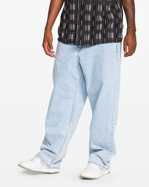 Wearing Volcom Men's Billow Denim Pant in light blue, paired with a black and white patterned short-sleeve shirt and white sneakers, a person stands against a plain white background. Only the lower body and part of the torso are visible.