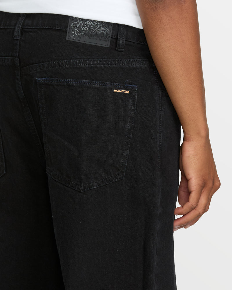 A person wearing black Men's Billow Denim jeans by Volcom is shown from the back, with their right arm at their side. The loose fit jeans feature a visible back pocket with a small Volcom logo tag.