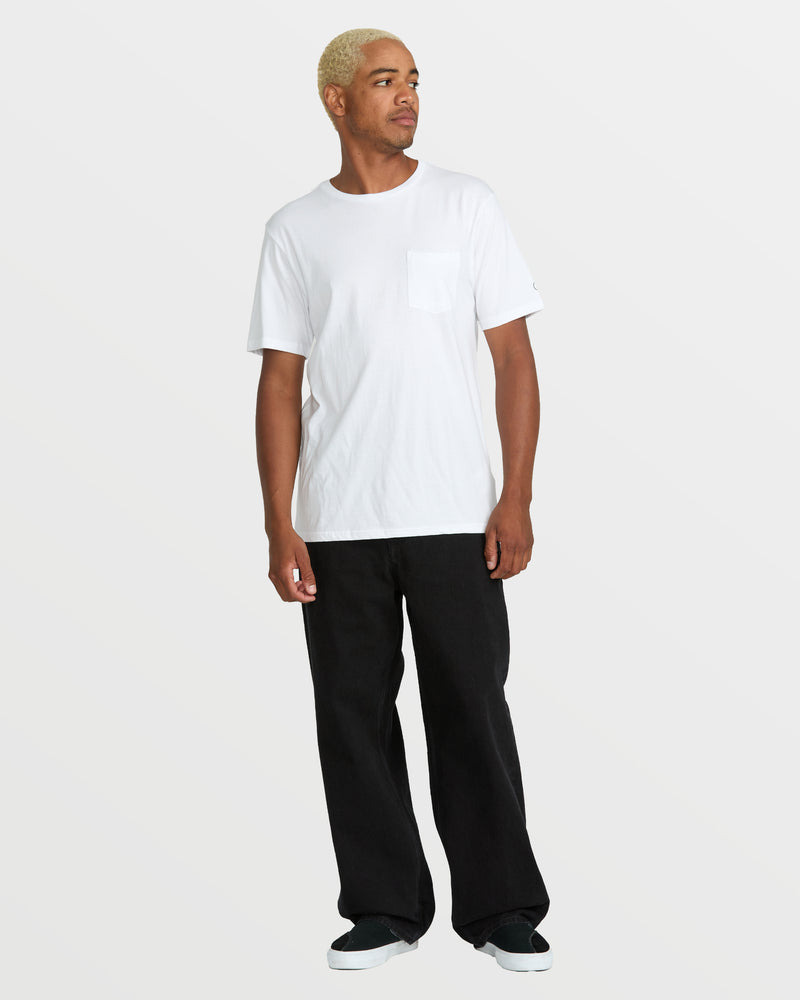 A person with short blond hair wears a white short-sleeve pocket t-shirt, Volcom Men's Billow Denim jeans in a loose fit, and black slip-on shoes, standing against a plain light gray background and looking to the side.