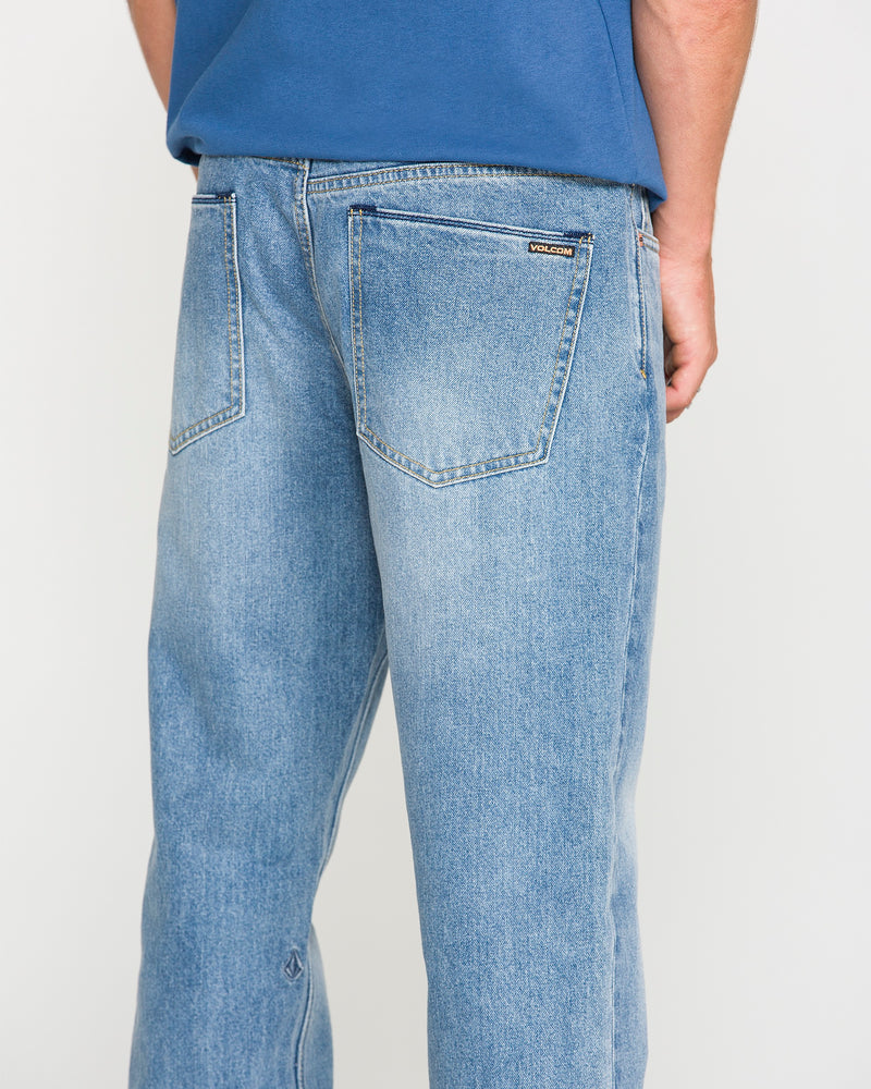 A person wearing Volcom Men's Solver Denim Pants in Freedom Blue and a blue shirt, shown from the back. The stretch denim jeans feature two back pockets and a small Volcom label on the right pocket.