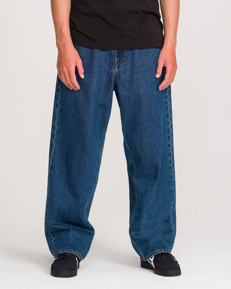 A person is shown from the shoulders down, standing against a plain white background, wearing Volcom’s Men’s Billow Denim Pants—loose-fitting, low-rise jeans in black—paired with black sneakers and a black t-shirt.
