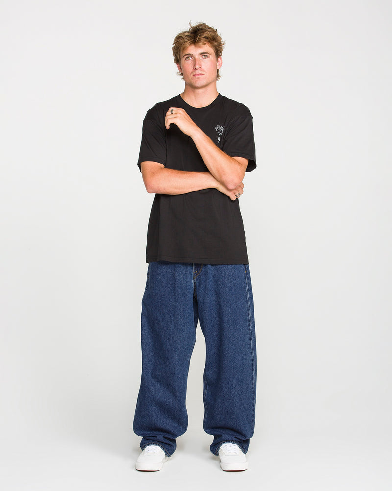 A young man stands against a plain background, wearing a black t-shirt, white sneakers, and Volcom Men's Billow Denim Pants in a loose fit dark indigo wash. One hand is raised near his face as he maintains a neutral expression.