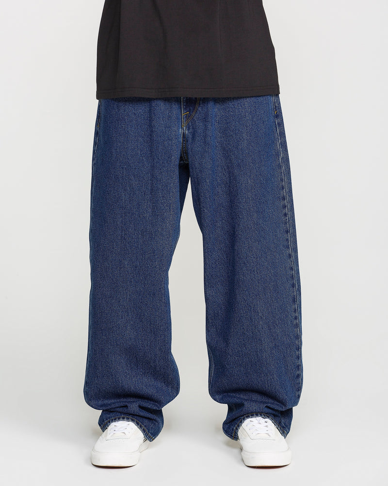 A person is shown from the waist down, wearing Volcom Men's Billow Denim Pants in a loose fit dark indigo wash, paired with a black shirt and white sneakers, standing against a plain white background.
