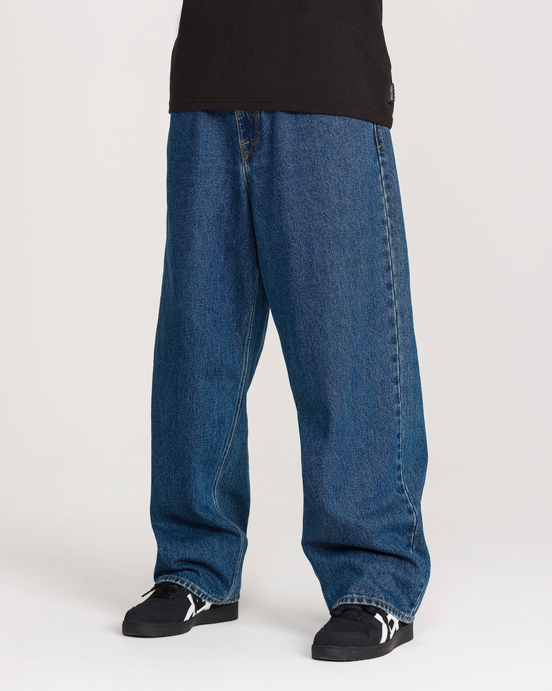 A person wearing a black shirt, Volcom Men’s Billow Denim Pants in a loose baggy blue fit, and black sneakers with white accents, standing against a plain light background.