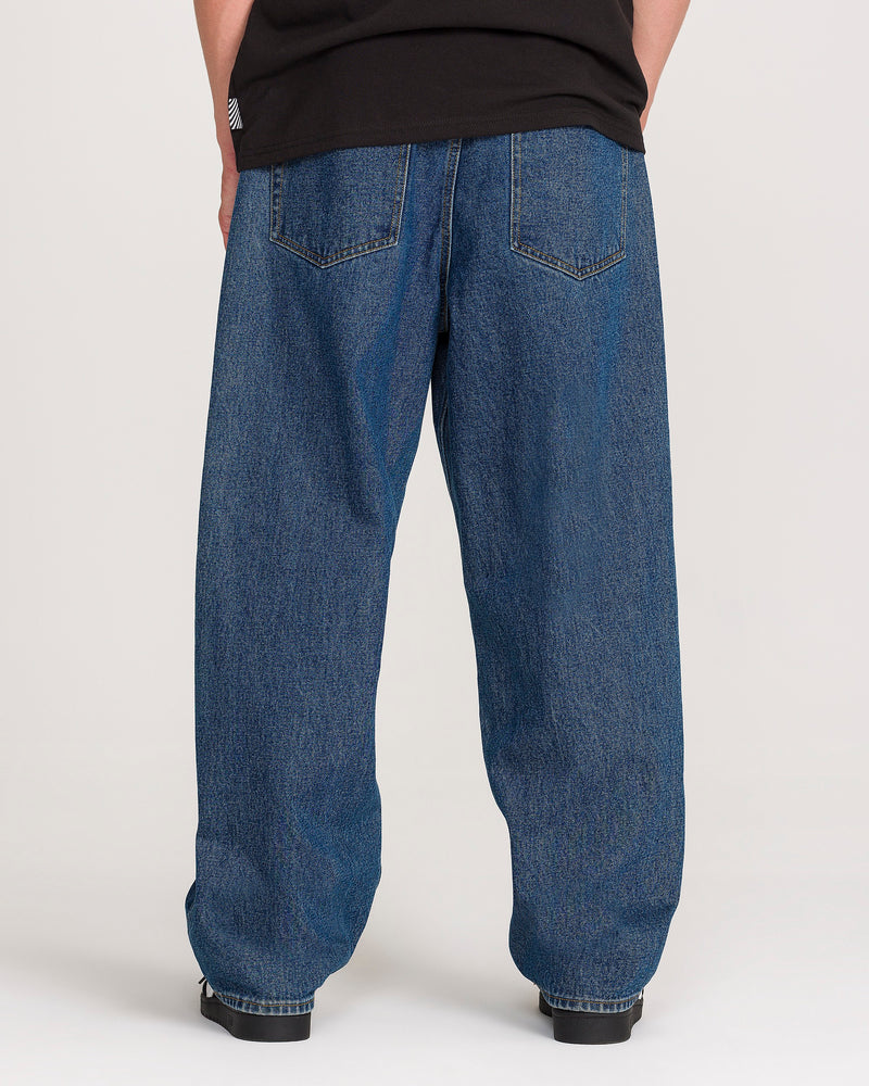 A person is seen from behind against a plain background, wearing Volcom Men’s Billow Denim Pants in dark indigo paired with black shoes.