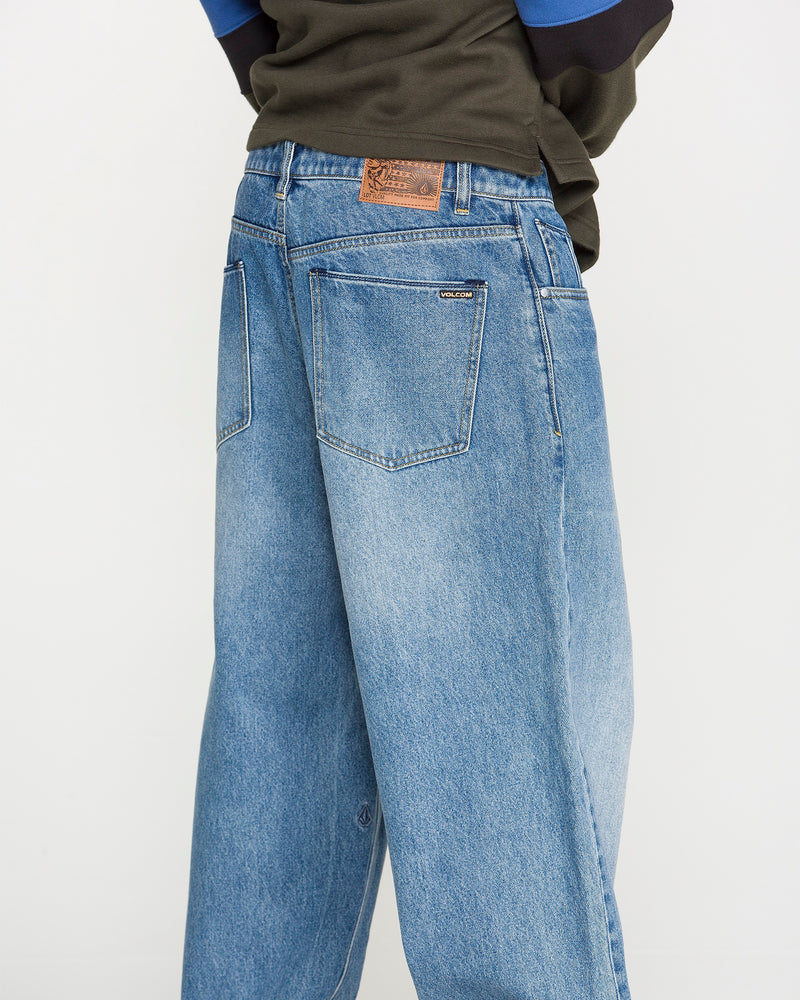 A person wearing Volcom Men's Chillow Denim Pants, featuring a relaxed fit in blue denim with visible back pockets and a brown patch on the waistband, is photographed from behind against a plain background.