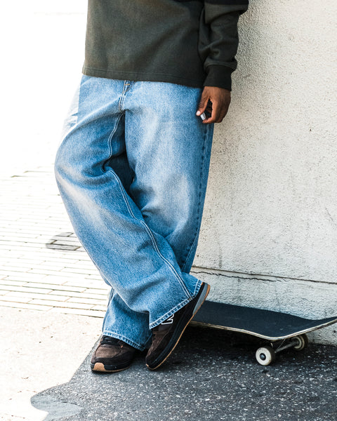 Wearing Volcom’s Men’s Chillow Denim Pants in washed indigo and a dark sweatshirt, the person leans against a white wall with just their lower body visible, a skateboard resting nearby.