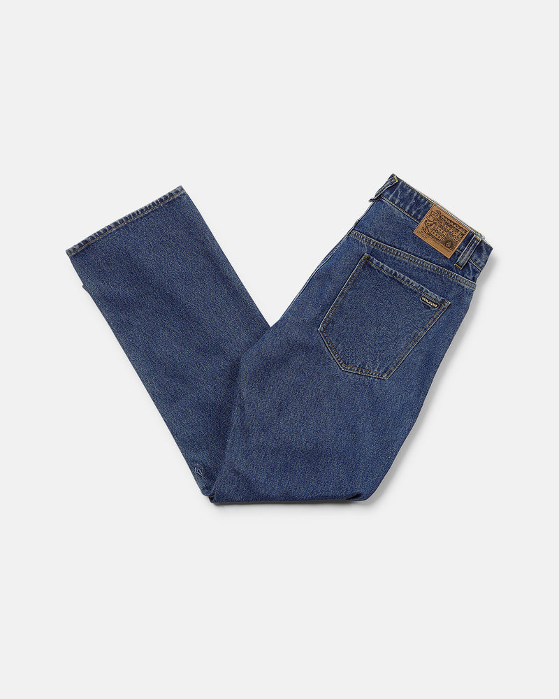 A pair of Volcom Men’s Pipe Dreams Denim Pants in dark indigo is folded neatly against a white background, displaying the back pocket and a brown leather patch on the waistband. Crafted from rigid 100% cotton denim, these jeans offer classic style and everyday comfort in a relaxed straight fit.