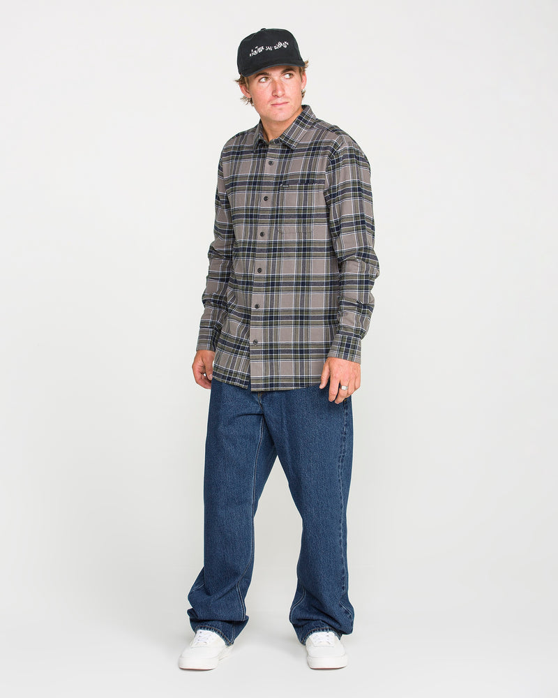 A man wearing a black cap, plaid button-up shirt, Volcom Men's Pipe Dreams Denim Pants in dark indigo, and white sneakers stands against a plain white background, looking slightly to his left.