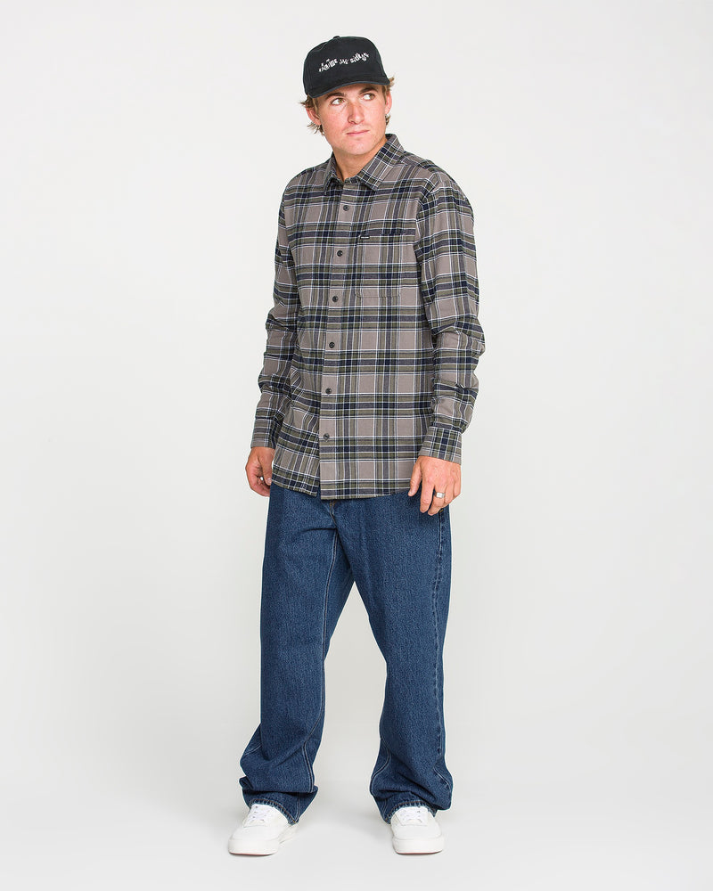 A man wearing a black cap, a gray plaid button-up shirt, white sneakers, and Volcom Men’s Pipe Dreams Denim Pants—relaxed straight fit jeans crafted from 100% cotton denim—stands against a plain light background.