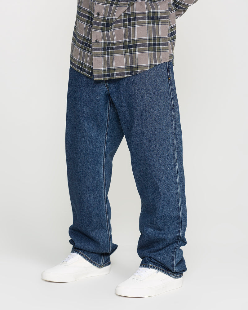 A person wearing Volcom Men's Pipe Dreams Denim Pants in a relaxed straight fit dark indigo, paired with a gray plaid shirt and white sneakers, stands against a plain light background. Only the lower half of the body is visible.