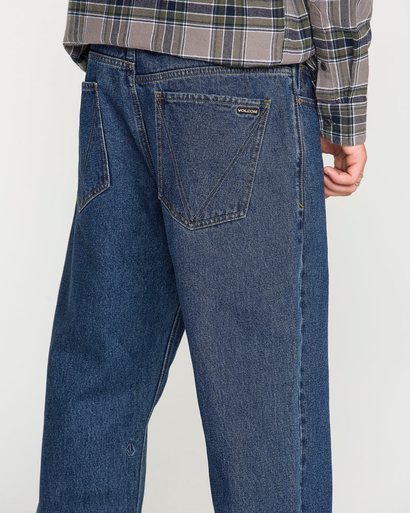 A person is shown from the back wearing Volcom Men's Pipe Dreams Denim Pants in a relaxed straight fit dark indigo denim, paired with a gray plaid shirt. One hand is partially visible and the jeans feature large back pockets and a small Volcom brand tag.