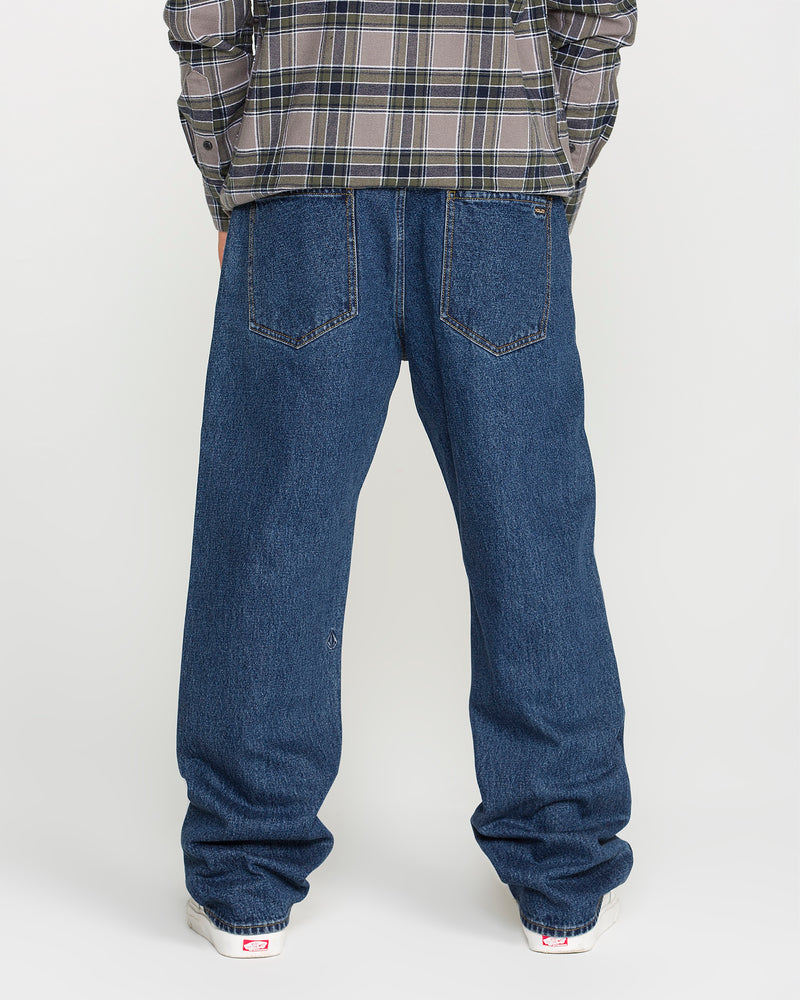 A person wearing a gray plaid shirt, Volcom Men’s Pipe Dreams Denim Pants in classic blue, and white sneakers is standing with their back to the camera against a plain light-colored background.