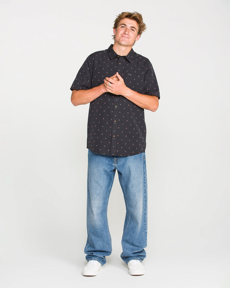 A person stands against a white background, wearing a black short-sleeve button-up shirt with a small pattern, Volcom Men’s Pipe Dreams Denim Pants in a relaxed straight fit vintage-inspired blue denim, and white sneakers, with hands clasped over their chest and a slight smile.