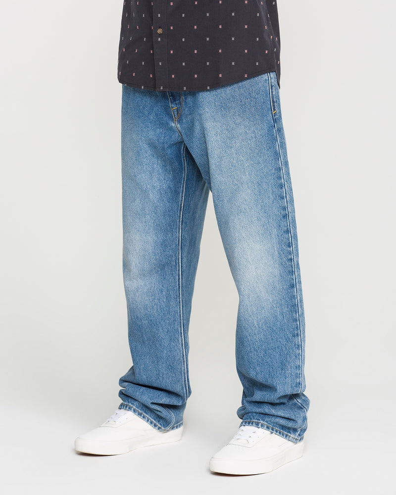 A person wearing Volcom Men’s Pipe Dreams Denim Pants—loose-fitting, vintage-inspired blue jeans with a relaxed straight fit—paired with a black shirt featuring small white and red patterns and white sneakers, standing against a plain light background.