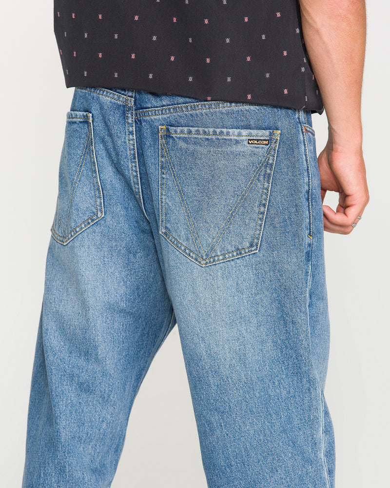 A person is shown from the waist down wearing Volcom Men's Pipe Dreams Denim Pants in Euro Light Blue—a vintage-inspired, relaxed straight fit with loose-fitting blue jeans, oversized back pockets, and a small “Volcom” label on one pocket. The jeans are paired with a black patterned shirt.