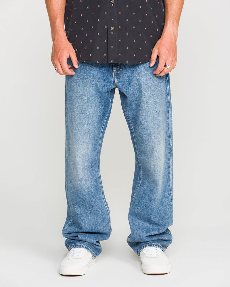 A person wearing a black patterned shirt, Volcom Men's Pipe Dreams Denim Pants in a relaxed straight fit, and white sneakers stands against a plain background. Only their legs and hands are visible.