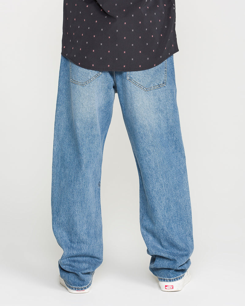 A person stands with their back to the camera and feet slightly apart on a plain background, wearing a black shirt with small red dots paired with Volcom’s Men’s Pipe Dreams Denim Pants in a relaxed straight fit and vintage-inspired light blue wash.