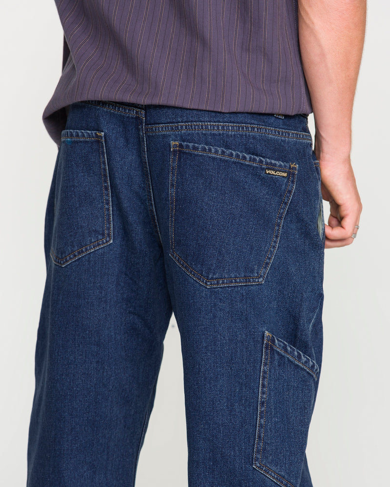 A person wearing Volcom Men's Kinkade Utility Denim Pants in dark blue indigo, featuring visible back and side pockets, paired with a purple striped shirt. The photo is taken from behind, highlighting the straight-leg utility denim pants and part of the shirt.