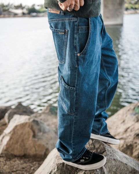 A person wearing Volcom Men’s Kinkade Utility Denim Pants and black sneakers stands on rocks by a body of water, with part of a bridge and trees visible in the background.