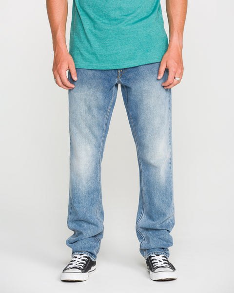 A person wearing a green t-shirt, Volcom Men's Vorta Denim Pants in a slim fit, and black-and-white sneakers stands with arms relaxed at their sides against a plain background.