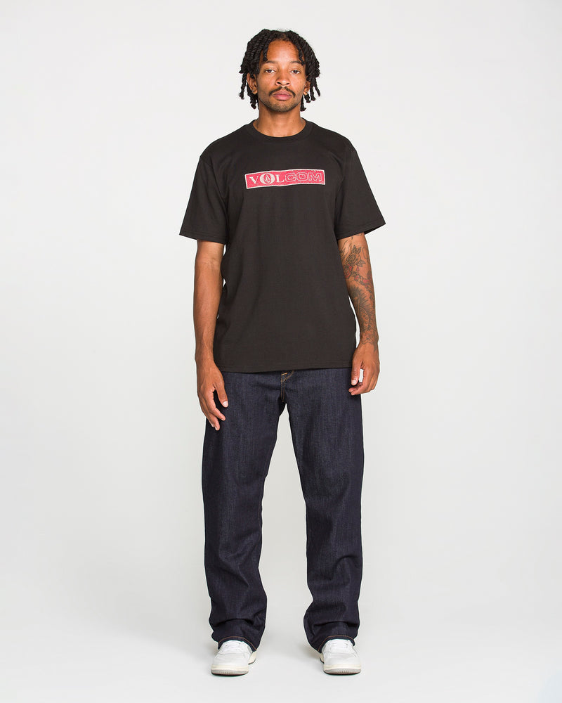 A man with short dreadlocks and tattoos on his left arm stands facing forward against a plain white background, wearing a black Volcom T-shirt, white shoes, and Volcom Men’s Pipe Dreams Denim Pants in indigo stretch denim with a relaxed straight leg fit.