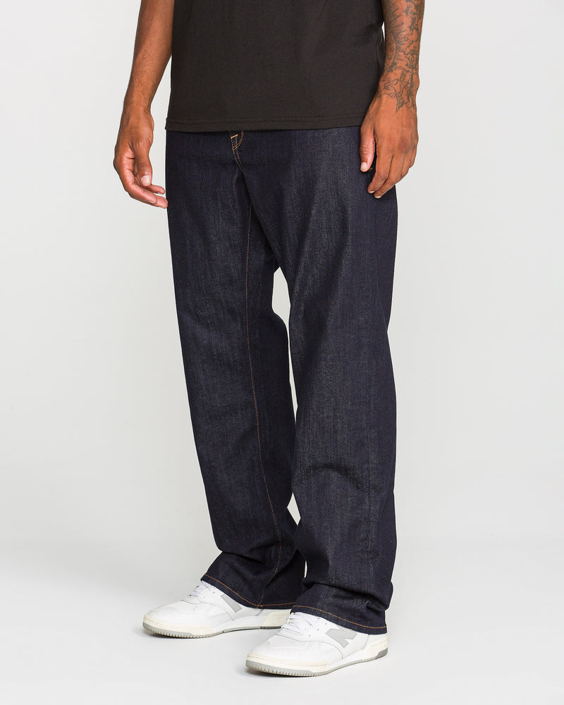 Wearing Volcom’s Men’s Pipe Dreams Denim Pants in indigo stretch denim, paired with a black t-shirt and white sneakers, an individual stands against a plain background. Only the lower half of the body is visible.