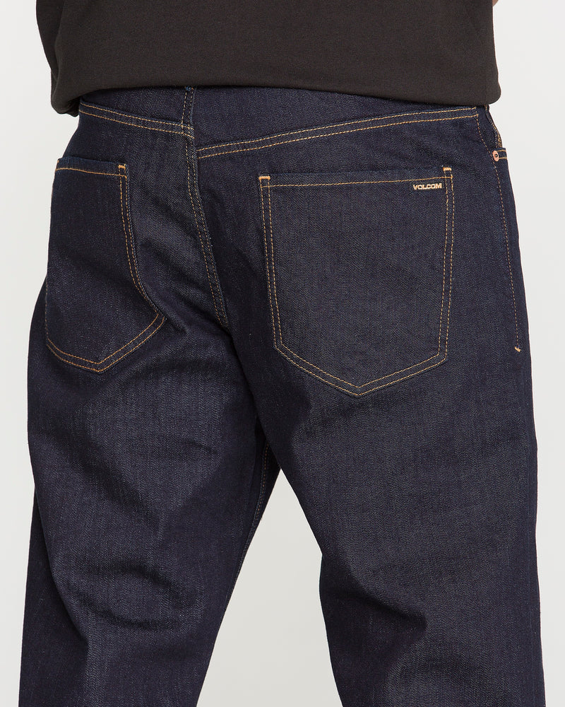 A person is seen from the back wearing Volcom Men’s Pipe Dreams Denim Pants in dark blue indigo stretch denim with visible orange stitching. The relaxed straight leg jeans feature two back pockets and have the Volcom logo stitched above the right pocket. The person pairs them with a dark shirt.