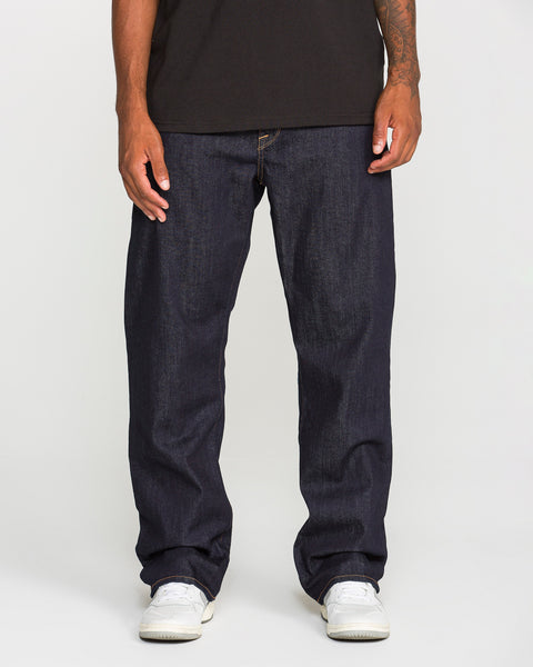 A person wearing a black t-shirt, Volcom Men’s Pipe Dreams Denim Pants in indigo stretch denim, and white sneakers stands against a plain white background. The photo is cropped at the shoulders.