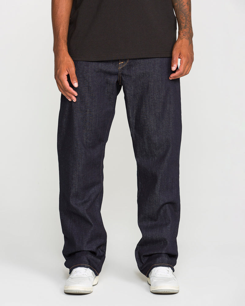 A person wearing a black t-shirt, Volcom Men’s Pipe Dreams Denim Pants in indigo stretch denim, and white sneakers stands against a plain white background. The photo is cropped at the shoulders.