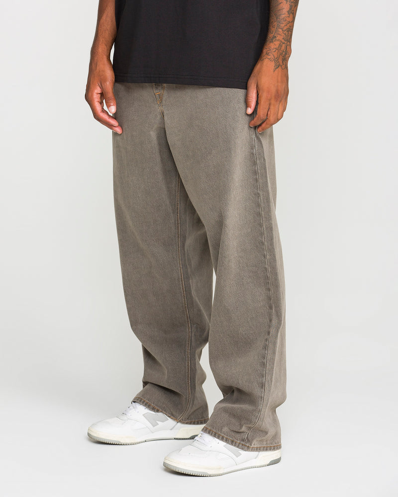 A person is shown from the waist down, standing against a plain white background and wearing Volcom Men's Featured Artist Antonio Carrau Reverb Pants in overdyed black cotton. The loose-fitting, low-rise silhouette of the pants is highlighted, paired with a black shirt and white sneakers.