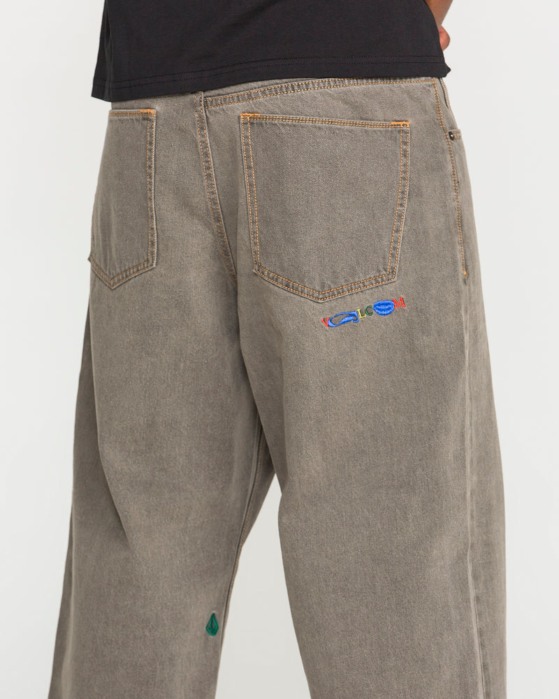 A person is seen wearing the Volcom Men's Featured Artist Antonio Carrau Reverb Pants—loose, gray-toned jeans with visible orange stitching, a vibrant embroidered motif near the right back pocket, and a small green accent by the left hem. Their upper body is partly shown in a black shirt.