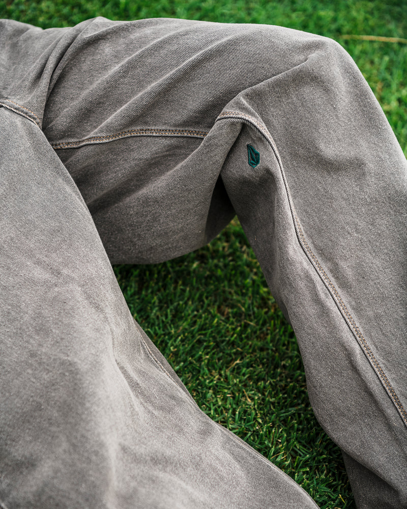 A close-up shows a person wearing Volcom Men’s Featured Artist Antonio Carrau Reverb Pants in gray, seated on green grass. A small dark green leaf-shaped logo is visible on the seam of the pants.