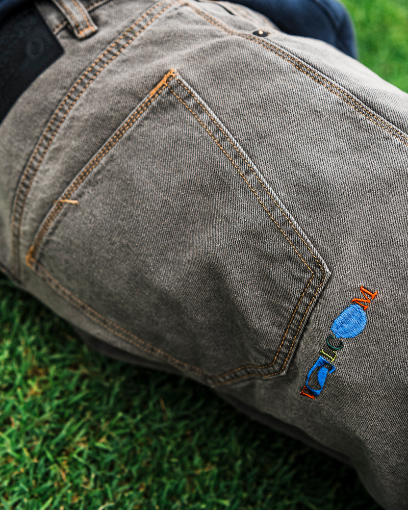 A close-up of the Volcom Men’s Featured Artist Antonio Carrau Reverb Pants in gray denim showcases orange stitching and colorful embroidery near the pocket, as part of a person lies on green grass. The low-rise fit highlights their casual style, while subtle black cotton detailing adds a modern touch.
