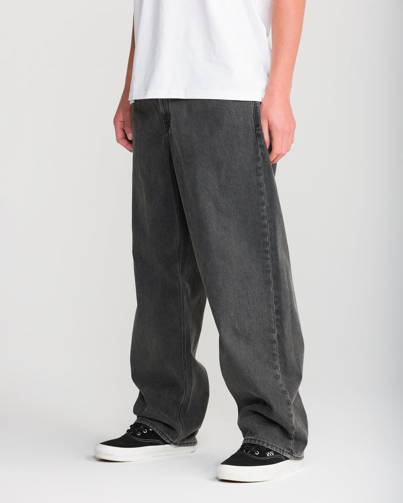 Person wearing a white t-shirt, Volcom Men’s Billow Denim in dark gray, and black-and-white sneakers stands against a plain light background; only the lower half is visible.