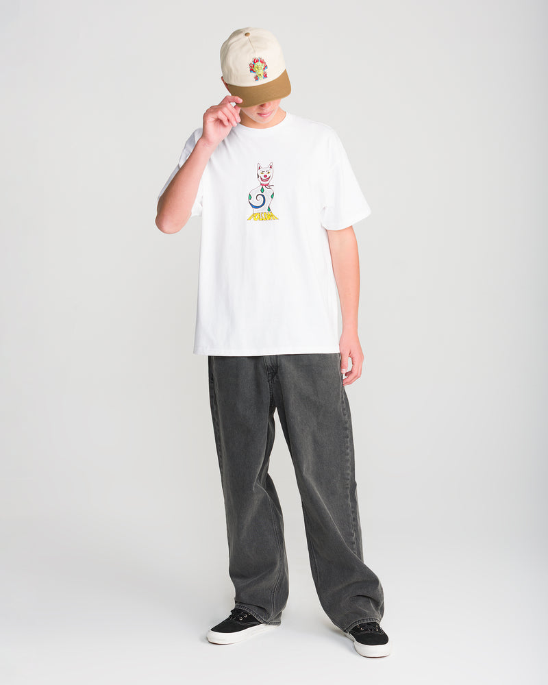 A person in a white tee with a colorful cat graphic, Men’s Billow Denim by Volcom, black sneakers, and a beige cap stands against a plain background, tipping their cap and looking slightly down.