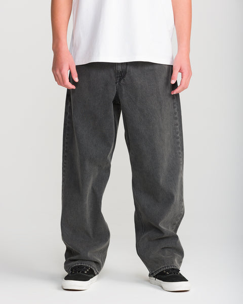 Person wearing Volcom Men’s Billow Denim in black, a white T-shirt, and black-and-white sneakers, standing against a plain light background. The image shows only the lower torso, legs, and hands.