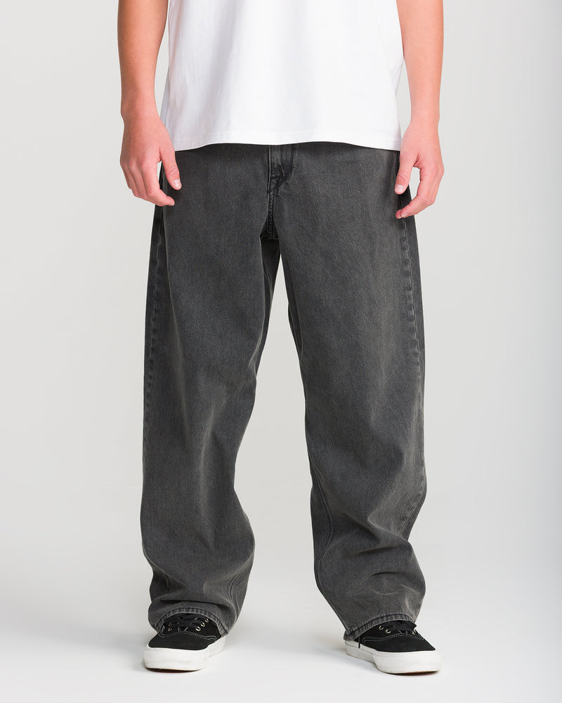 Person wearing Volcom Men’s Billow Denim in black, a white T-shirt, and black-and-white sneakers, standing against a plain light background. The image shows only the lower torso, legs, and hands.