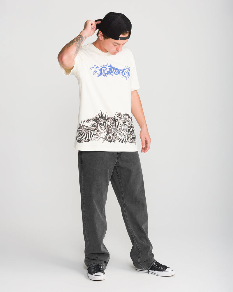 A person wearing a graphic t-shirt, Volcom Men’s Pipe Dreams Denim in black, and black sneakers stands while adjusting a baseball cap.