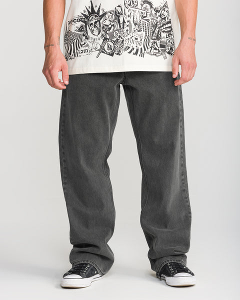 A person wearing Volcom Men’s Pipe Dreams Denim in charcoal grey with a loose, relaxed straight-fit and black sneakers.