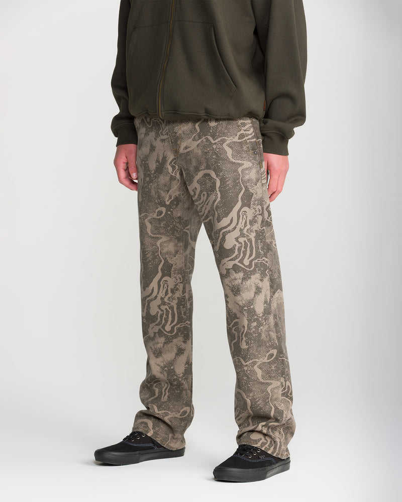 A person stands against a plain background wearing Volcom Men’s Kinkade Utility Denim, a dark green zip-up jacket, and black shoes. Their face is not visible in the image.