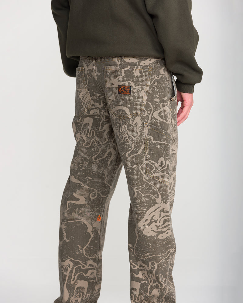 Person wearing an olive green jacket and Volcom Men’s Kinkade Utility Denim in grayish camo with a straight leg fit, orange logo, and brown patch by the pocket. Background is plain and light-colored.