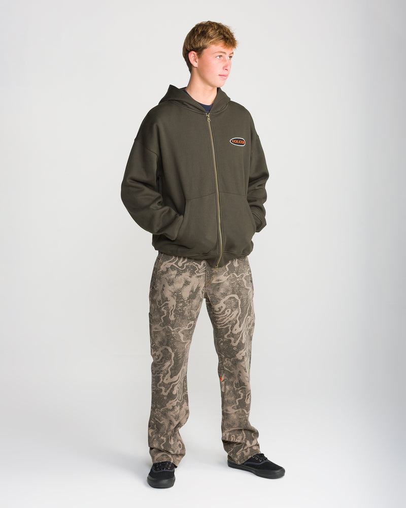 A young man with short light brown hair stands on a white background, wearing a dark zip-up hoodie with hands in pockets and Volcom Men’s Kinkade Utility Denim featuring dragon designs.