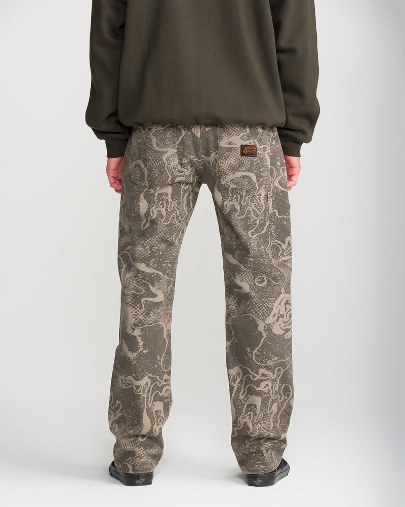 Person stands with their back to the camera, wearing Volcom Men’s Kinkade Utility Denim in a brown and gray camo pattern with a dark long-sleeve shirt against a light background.