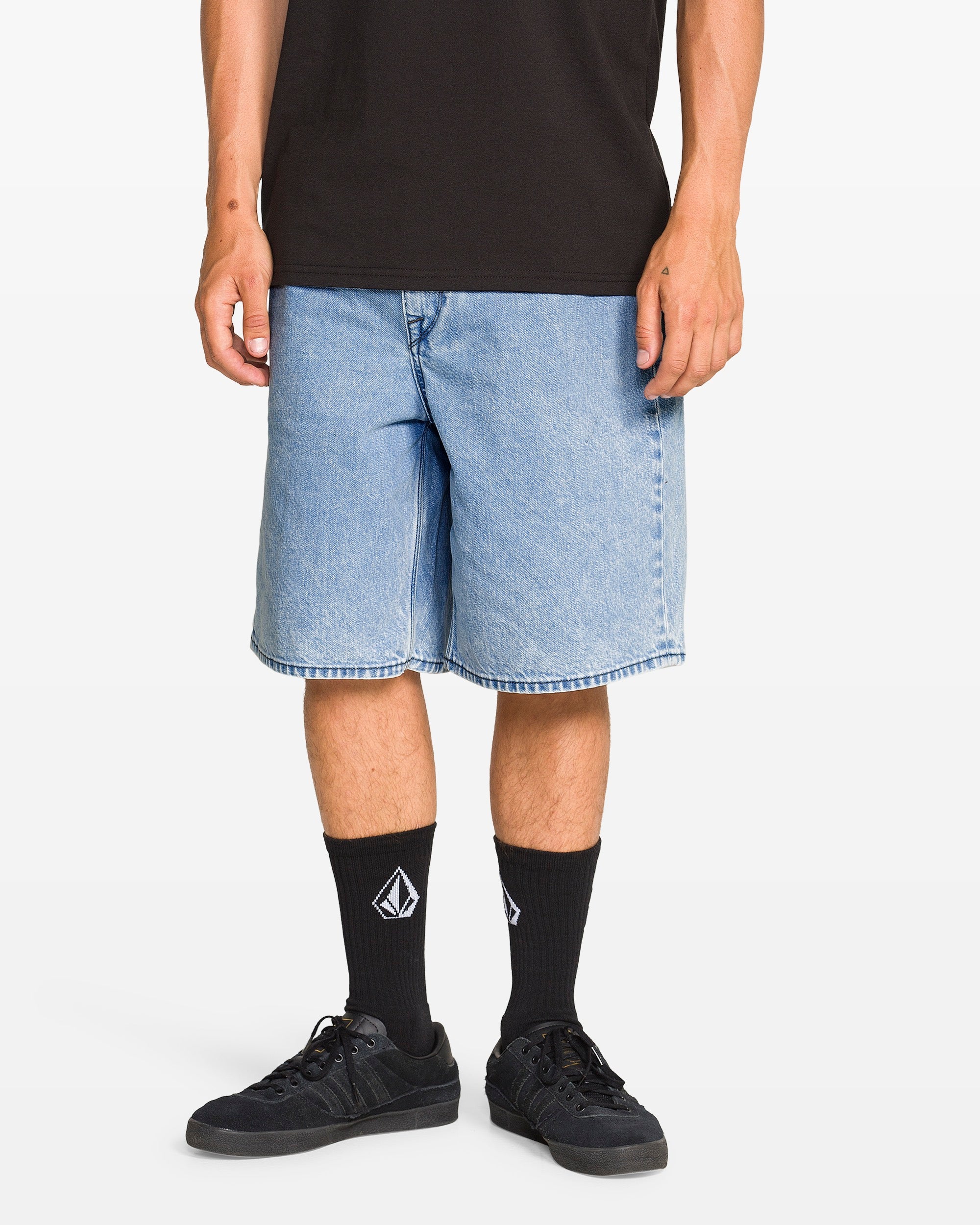 A person stands against a white background wearing Volcom Men's Billow Denim Shorts in a baggy, light blue fit made from recycled cotton, paired with a black t-shirt, black sneakers, and black socks with a white geometric logo. Face is not visible.