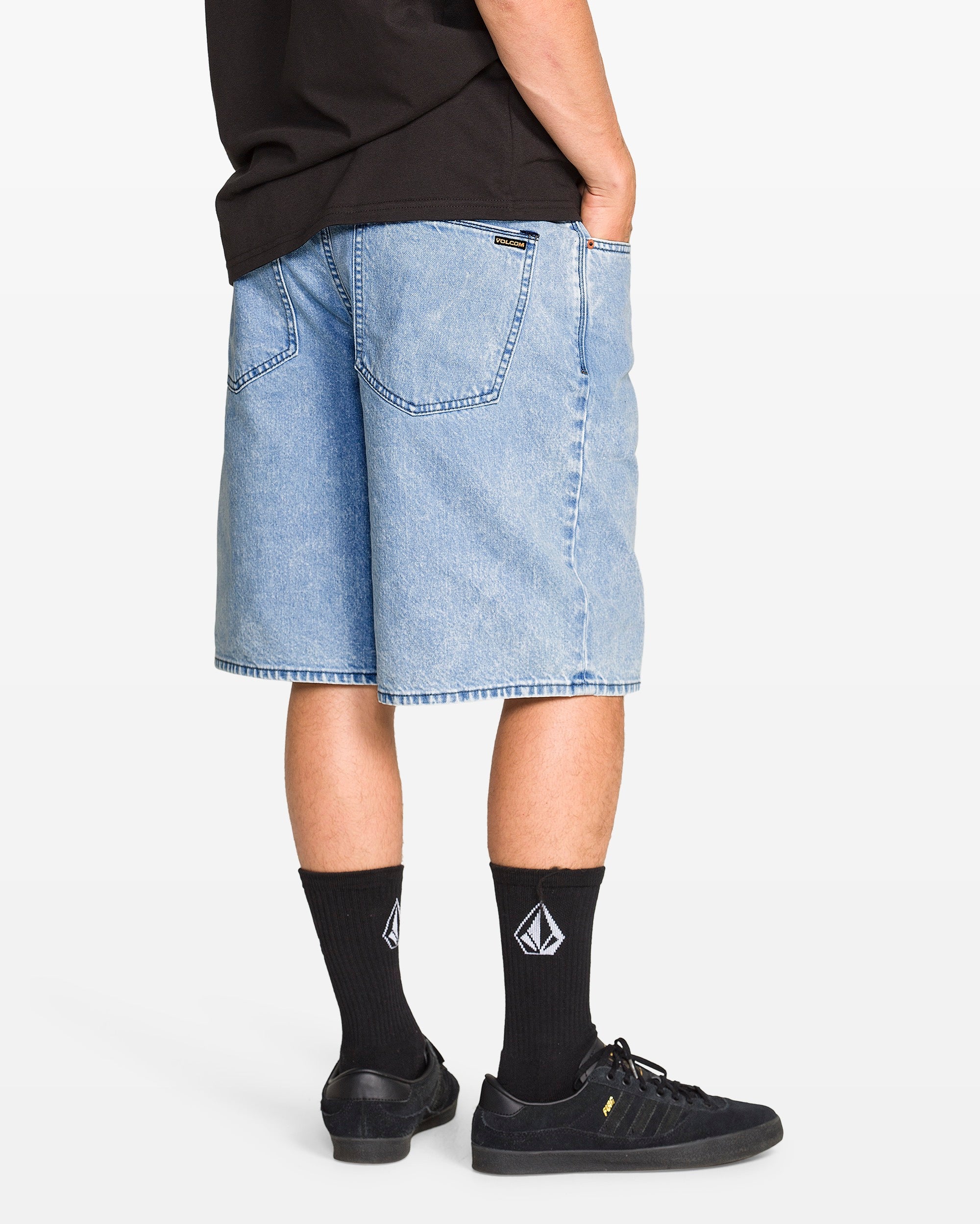 A person stands with hands in pockets, facing away from the camera against a plain background, wearing Volcom Men's Billow Denim Shorts made from recycled cotton, a black T-shirt, black socks with white logo, and black sneakers.